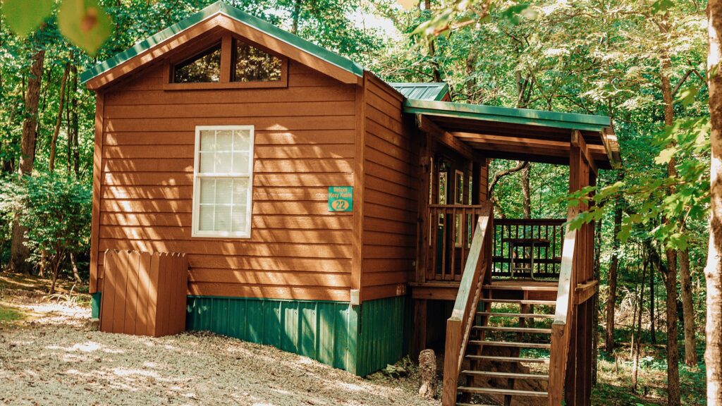 Wood cabin with a green metal roof and a covered porch with steps, tucked into shady woods