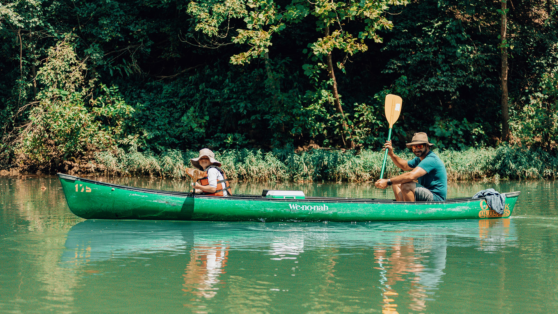 Two paddlers in a green canoe on the calm, tree-lined Meramec River