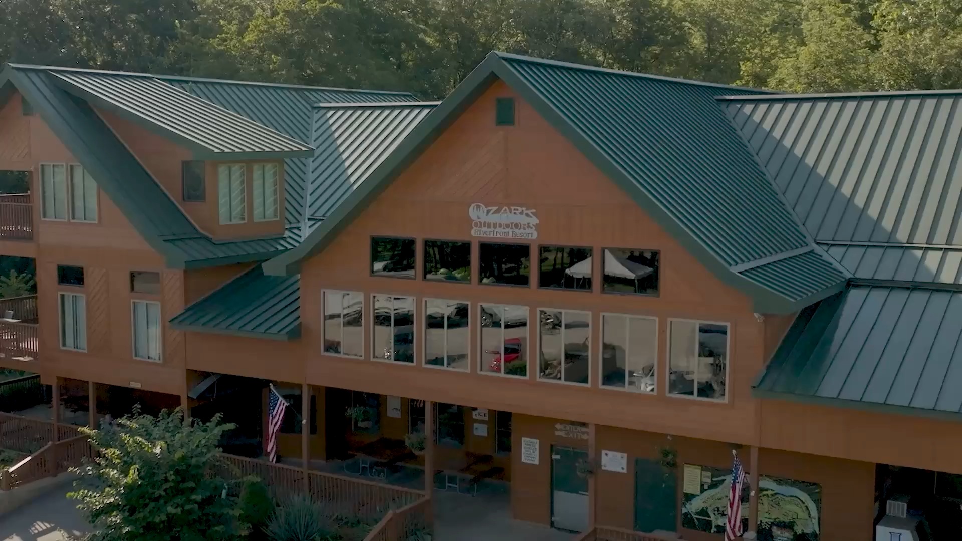 Aerial view of the Ozark Outdoors Riverfront Resort main building, with cedar siding and green metal roofs, backed by trees.