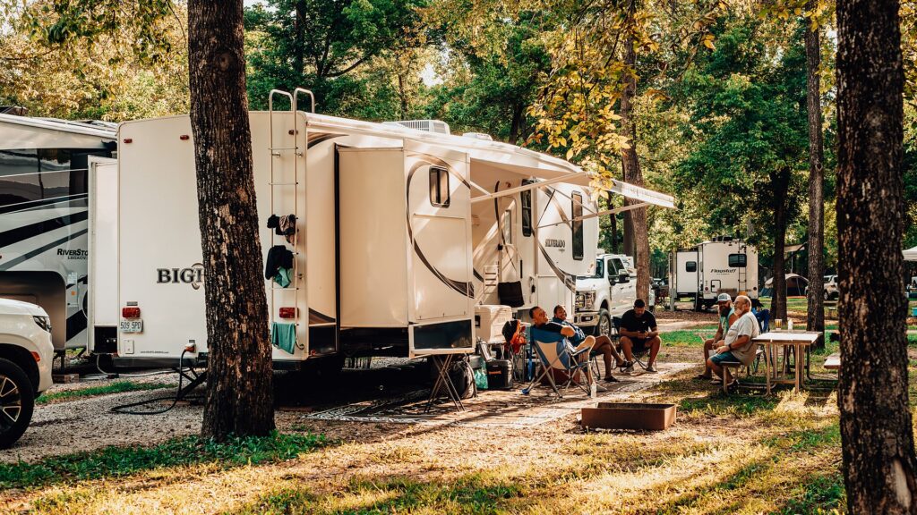 Campers relaxing in camp chairs beside a fifth-wheel RV on a shaded, tree-lined RV site