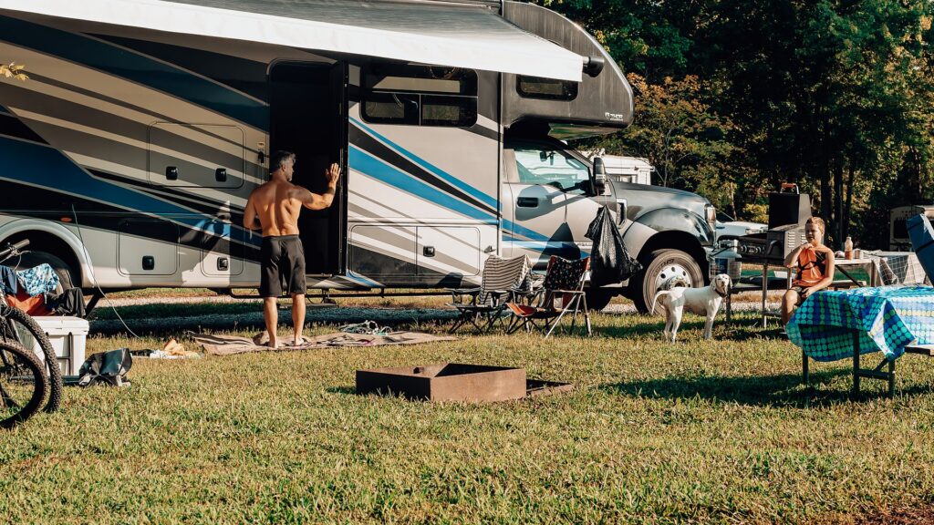 Family relaxing outside their Class C motorhome at a grassy RV site with a fire ring and their dog