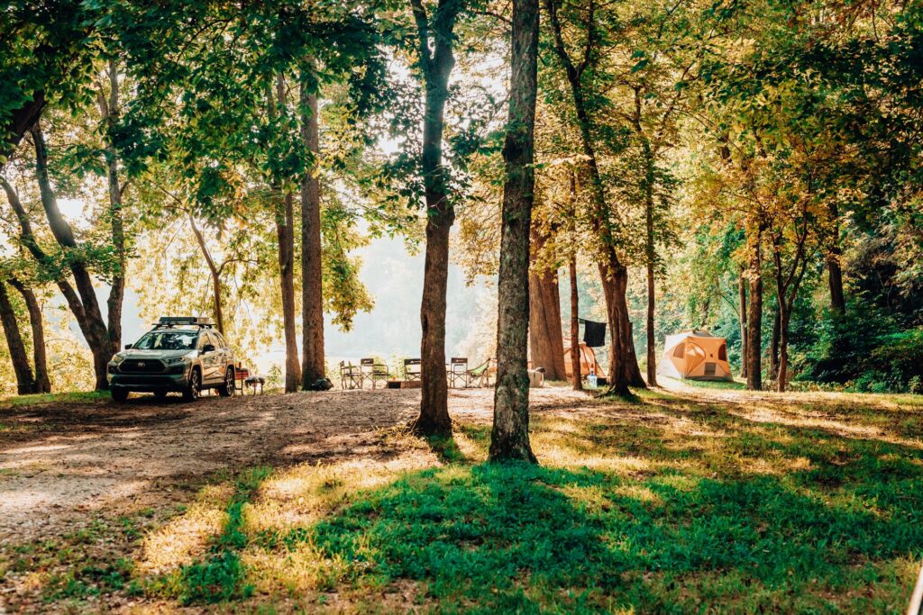 Riverside campsite at golden hour with an SUV, camp chairs and two tents under tall shade trees