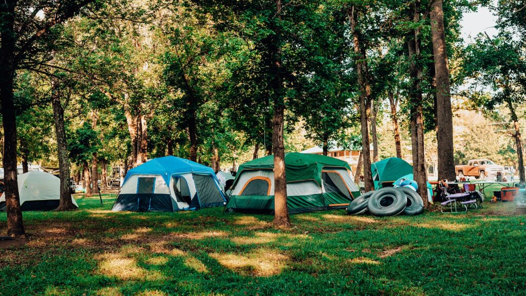 Row of dome tents pitched on shaded grass among tall trees, with river tubes stacked nearby