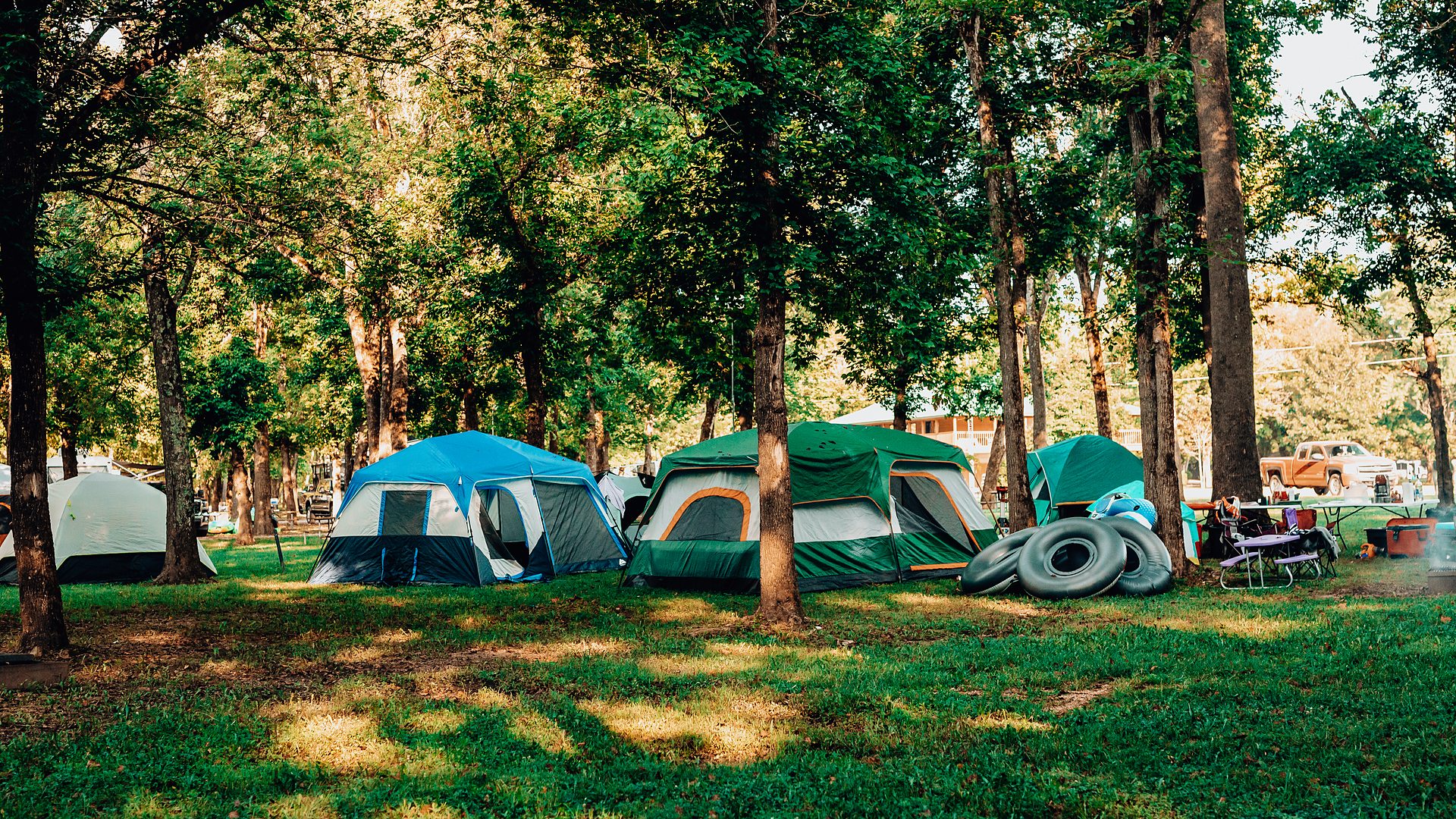 Row of dome tents pitched on shaded grass among tall trees, with river tubes stacked nearby